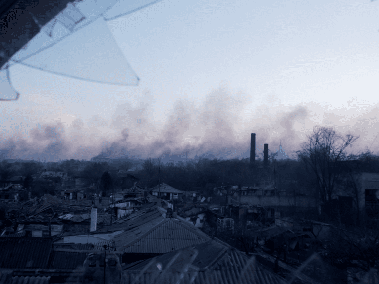 Zu sehen ist ein zerstörtes Dorf, über dem Rauchwolken schweben.
